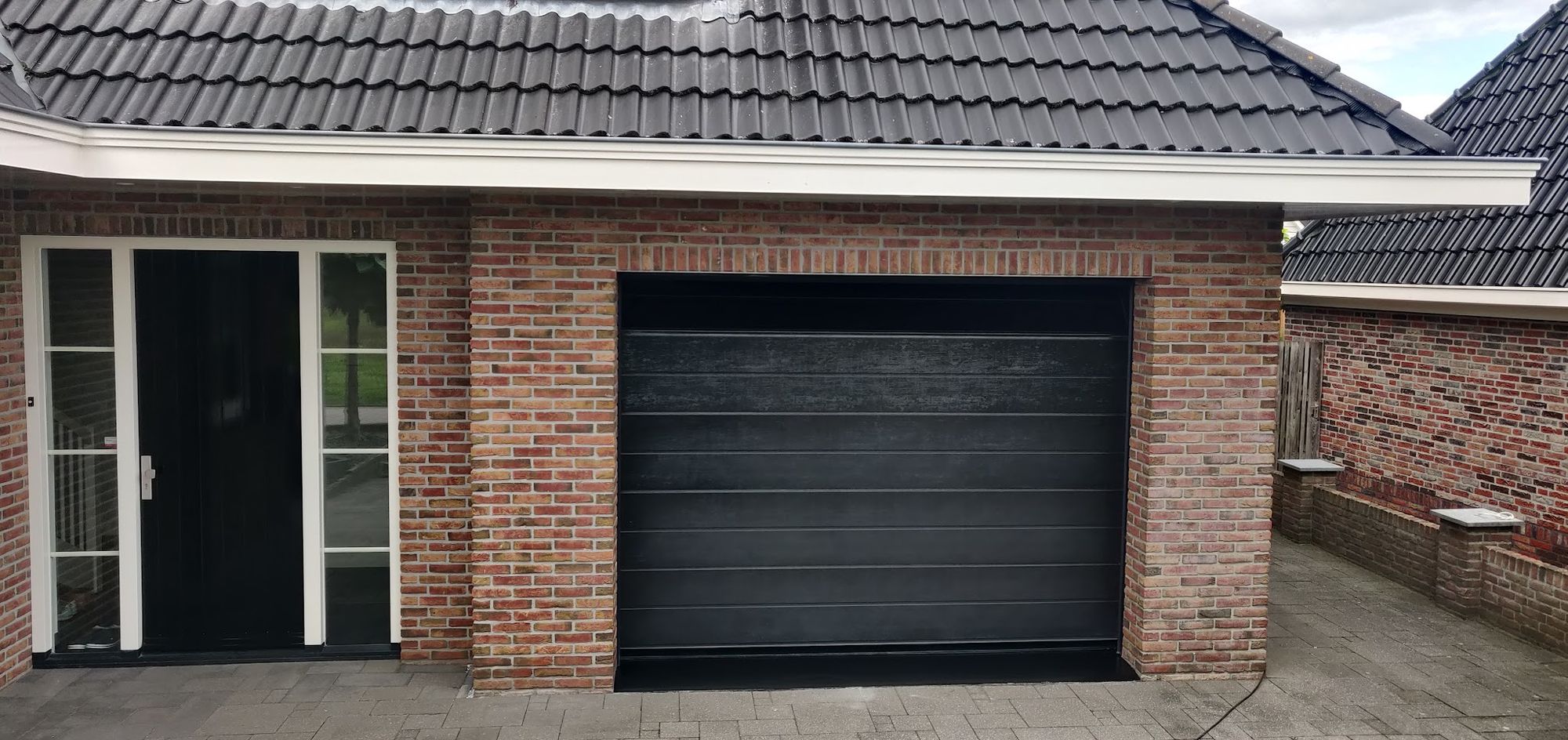 Een bakstenen garage met zwarte sectionaaldeur, witte kozijnen en een zwarte voordeur