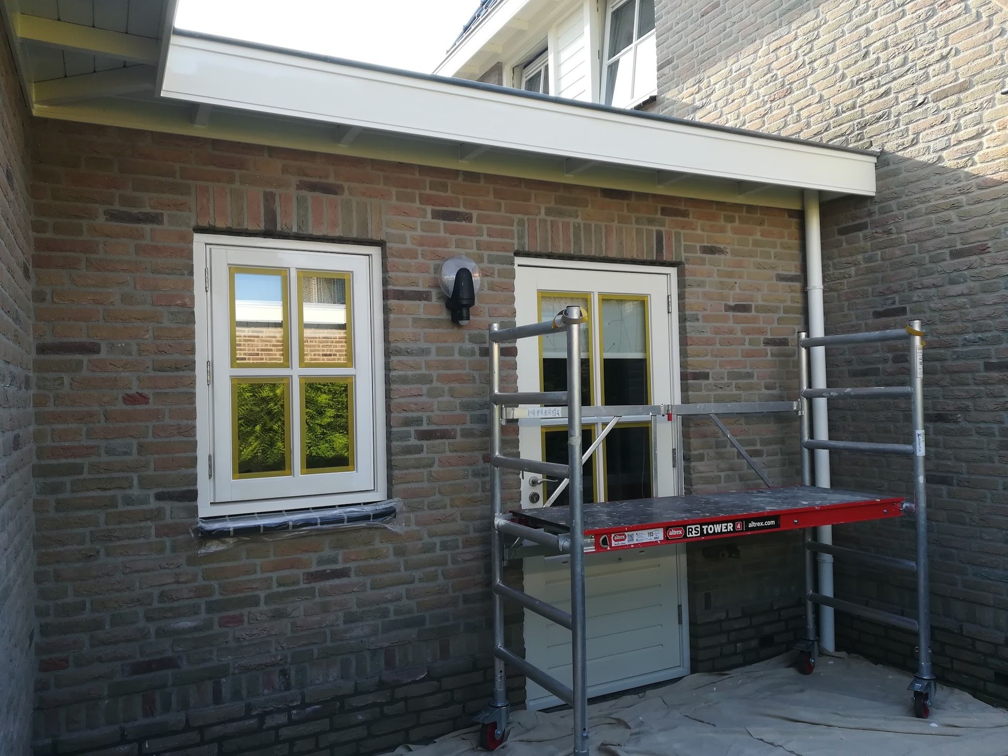 Crème geschilderde kozijnen en een deur in een bakstenen gevel, met een rolsteiger ervoor tijdens het werk