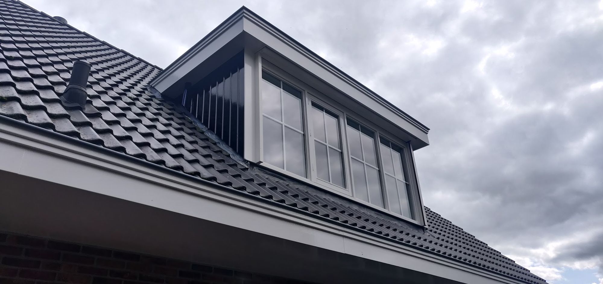 Een dakkapel met donkere beplating en witte kozijnen tegen een wolkenlucht